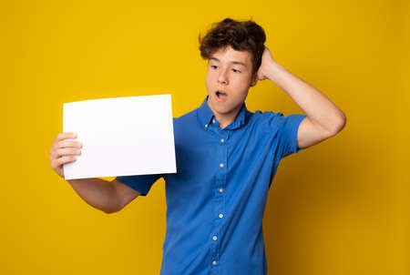 Beautiful european schoolboy in blue shirt posing with blank white paper against yellow background.の写真素材