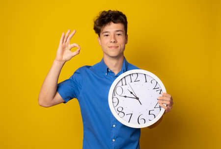 Portrait of caucasian teen boy with clock. Funny teenager showing clock, looking at camera. Child back to school, isolated on yellow. Education and time concept.の写真素材