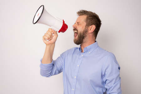 Portrait of a handsome young businessman wearing blue shirt standing isolated over white background, holding loudspeaker, screamingの写真素材
