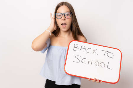 Amazed young girl wearing glasses holding white board over white background. Back to school concept.の写真素材
