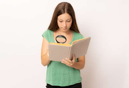 Young girl holding a book looking through a magnifying glass over white background.の写真素材