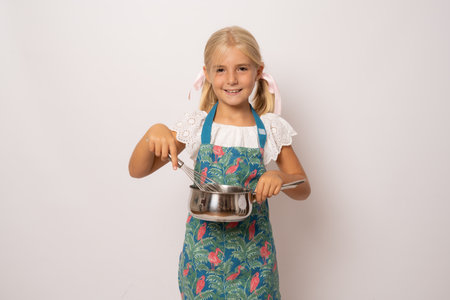 Young smiling girl wearing kitchen apron and holding an egg beater and a steel bowl over white backgroundの写真素材