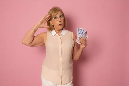 Shocked senior woman holding lots of cash isolated over pink background.の写真素材