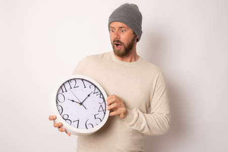 portrait of amazed young man in wool cap holding a clock with his hands over white backgroundの写真素材