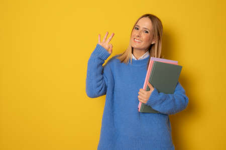 Portrait of happy young caucasian woman holding notebooks with okay sign isolated on yellow backgroundの写真素材