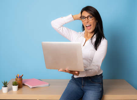 Attractive amazed business woman leaning on the table using laptop over blue background