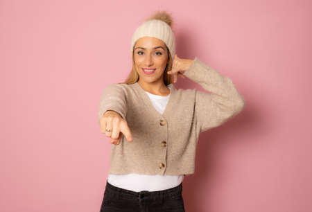 Young beautiful blonde woman wearing casual sweater over pink isolated background smiling doing talking on the telephone gesture and pointing to you. Call me.の写真素材