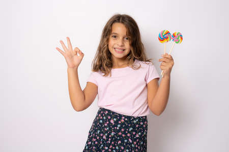 Beautiful little girl with lollipop and okay sign isolated over white background.の写真素材