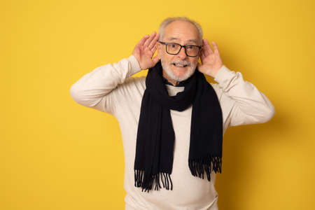 Senior man wearing winter clothing trying to hear both hands on ear gesture, curious for gossip. Hearing problem, deaf.の写真素材