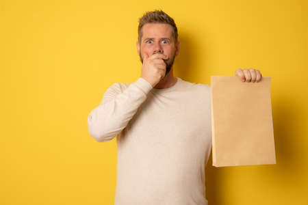Young amazed man showing brown clear blank craft paper takeaway bag mock up isolated on yellow background.の写真素材