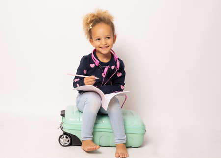 Cute little girl sits on a suitcase and writing in a notebook isolated over white background.の写真素材