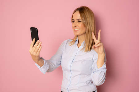 Happy woman holding mobile phone with victory sign isolated over pink background.の写真素材