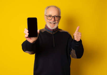 Lifestyle, technology and old people concept: Handsome old gray-haired man dressed in smart casual style using a smartphone and smiling over yellow backgroundの写真素材