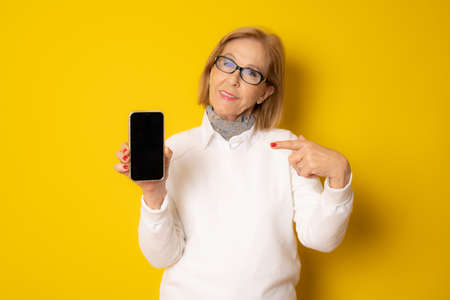 Mature smiling woman wearing white sweater showing smartphone isolated over yellow background.の写真素材
