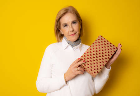 Attractive mature aged aged woman holding presents gift box feels happy studio isolated over yellow...