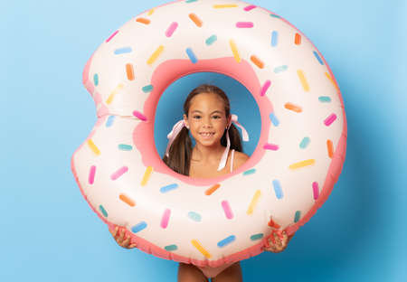 Cheerful little girl wearing swimsuit standing isolated over blue background, playing with inflatable ringの写真素材