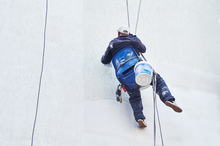 The builder repairs the facade of the building. The worker makes the thermal insulation of the outer wall of the apartment house. A man works in a hanging chair on ropesの写真素材