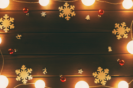 Frame holiday card. Bright Xmas garland along the perimeter of the frame. Christmas wooden decorations with snowflakes, horses, bells, red glass balls, on a background of brown old wooden boardsの写真素材