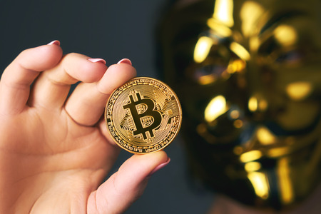 A gold coin bitcoin in the hand of a young woman on a dark background close-up and a blurred golden mask Anonymous. Crypto currencyの写真素材