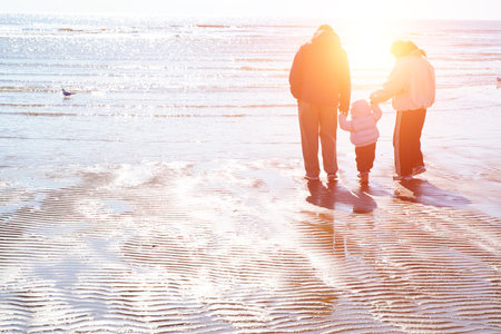 A family of three walks together on the wet, rippled beach at low tide, with the calm sea stretching...