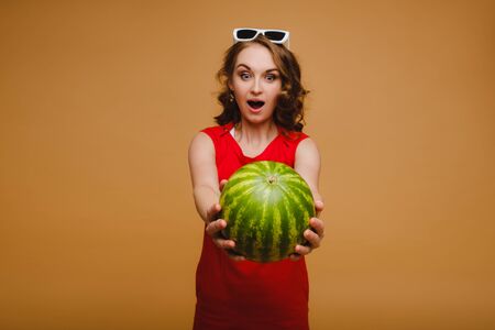 A beautiful girl in glasses and a red dress holds a watermelon in her handsの写真素材