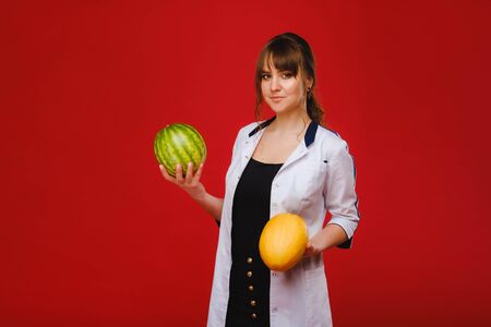 a female doctor nurse in a white coat with fruit in her hands poses on a red background, melon, watermelon,の写真素材
