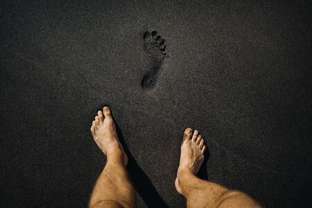 Close up of male footprints and feet walking on the volcanic black sand on the beach.の写真素材