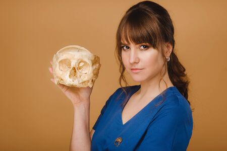 Cute young doctor girl holding a human skull, brown background behind the doctor