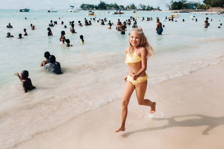 a little girl runs along a tropical beach with locals on the island of Mauritius.a girl on the beach of the Indian ocean and unrecognizable locals of the island of Mauritius.の写真素材