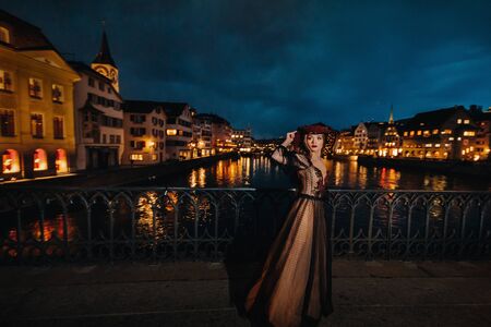 A stylish bride in a black wedding dress and red hat poses at night in the old city of Zurich....