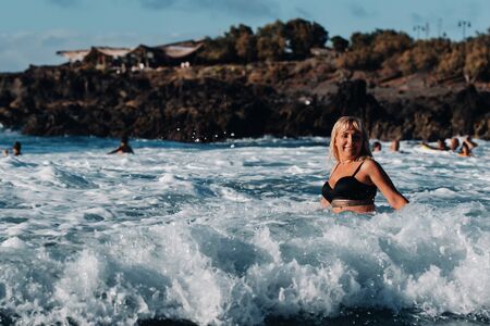 A girl with wet hair and a black swimsuit stands in the white foam of sea water on the island of Tenerife, around a wave with splashes of spray and water drops.Canary Islands.Spainの写真素材