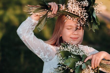 A beautiful nine-year-old blonde girl with long hair in a long white dress, holding a bouquet of lilies of the valley flowers, walking in nature in the Park.Summer, sunset.の写真素材