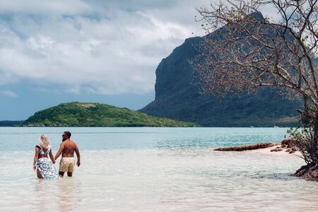 A girl in a swimsuit and a man in shorts stand in the ocean against the backdrop of mount Le Morne on the island of Mauritius.A couple in the water look into the distance of the ocean against the background of mount Le Morne .の写真素材