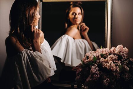 Beautiful girl in the mirror reflection at home. Girl before the holiday near the home mirror.A girl in a white dress with long hair is going to a party at home.の写真素材
