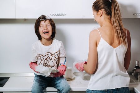 housewife mom in pink gloves washes dishes with her son by hand in the sink with detergent. A girl in white and a child with a cast cleans the house and washes dishes in homemade pink gloves.A child with a cast washes dishes and smiles.の写真素材