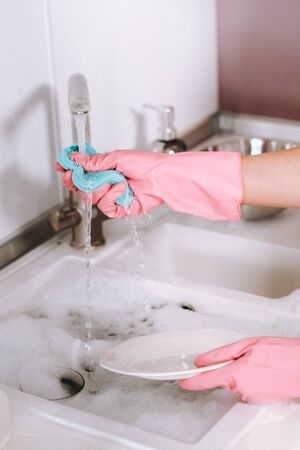 housewife girl in pink gloves washes dishes by hand in the sink with detergent. The girl cleans the house and washes dishes in gloves at homeの写真素材