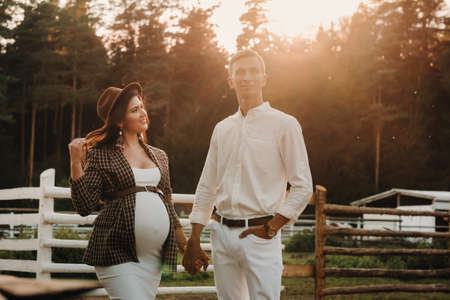 a pregnant girl in a hat and her husband in white clothes stand next to the horse corral.a stylish couple waiting for a child stand on the street near the horse corral.の写真素材