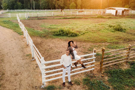 a pregnant girl in a hat and her husband in white clothes stand next to the horse corral.a stylish couple waiting for a child stand on the street near the horse corral.の写真素材