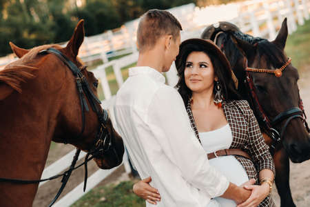 a pregnant girl in a hat and her husband in white clothes stand next to the horses near the horse corral.Stylish pregnant woman with a man with horses.Familyの写真素材
