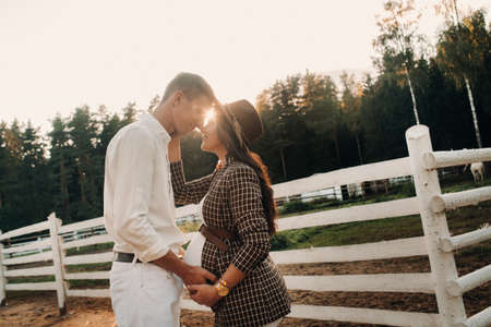 a pregnant girl in a hat and her husband in white clothes stand next to the horse corral.a stylish couple waiting for a child stand on the street near the horse corral.の写真素材