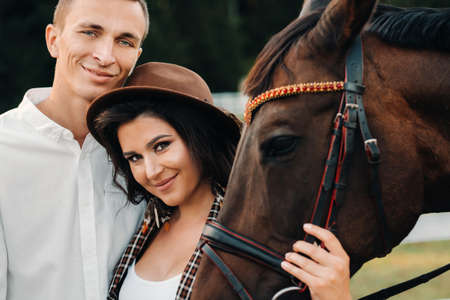 a pregnant girl in a hat and a man in white clothes stand next to horses near a white fence.Stylish pregnant woman with a man with horses.Married couple.の写真素材