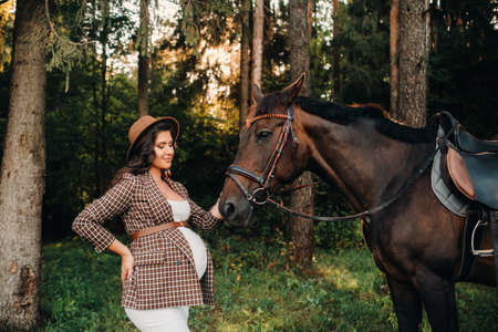 pregnant girl with a big belly in a hat next to horses in the forest in nature.A stylish pregnant woman in a white dress and brown jacket with horses.の写真素材
