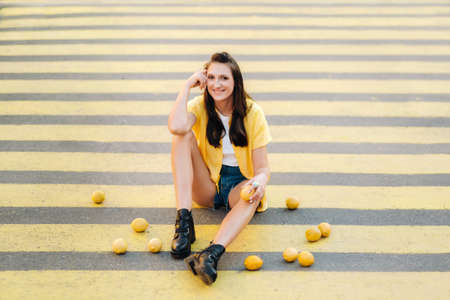 a creative girl in yellow clothes with lemons sits on a yellow pedestrian crossing in the city. The...