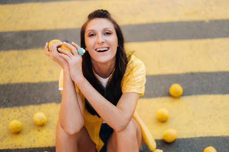 a creative girl in yellow clothes with lemons sits on a yellow pedestrian crossing in the city. The lemon mood.の写真素材