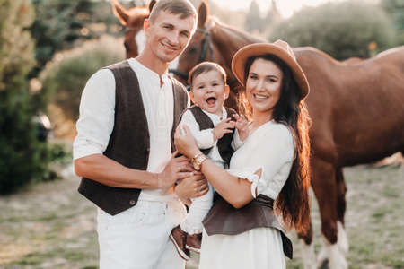A family in white clothes with their son stand near two beautiful horses in nature. A stylish couple with a child are photographed with horsesの写真素材