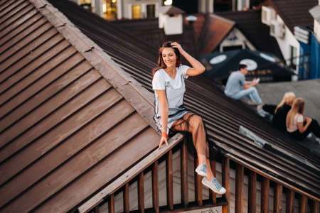 A stylish girl in a light dress and sneakers is sitting on the roof of a house in the evening in the city centerの写真素材