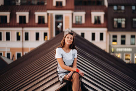 A stylish girl in a light dress and sneakers is sitting on the roof of a house in the evening in the city centerの写真素材