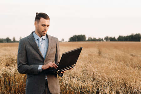 Portrait of a businessman in a suit holding a laptop in a field of wheat against the background of a windmill and the evening sky.の写真素材