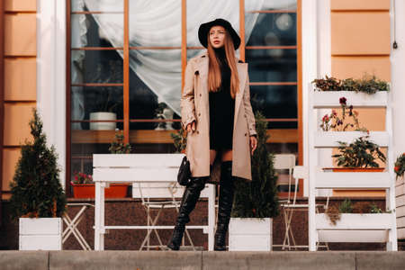 Stylish young woman in a beige coat in a black hat on a city street. Women's street fashion. Autumn...