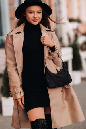Stylish young woman in a beige coat and black hat and with a black purse on a city street. Women's street fashion. Autumn clothing.Urban style.の写真素材